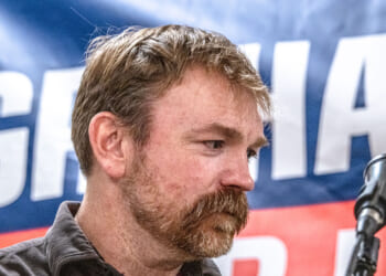 Democrat Graham Platner looks down, with a microphone beside him and his campaign banner partially seen in the background.