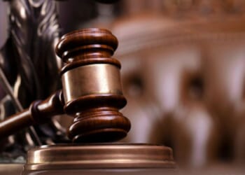 A gavel sits on a wooden block while a tufted leather seat and Lady Justice statuette sit behind.