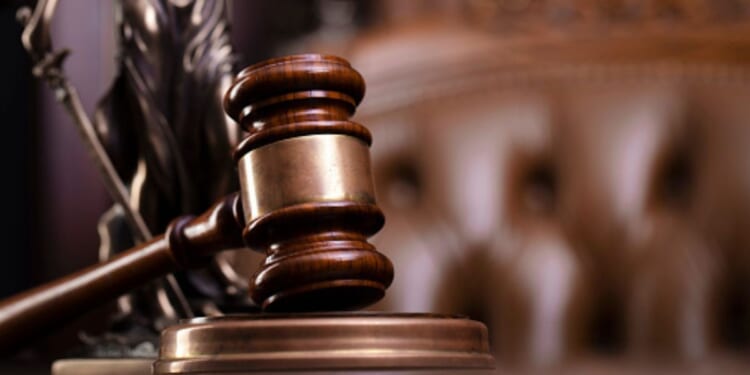 A gavel sits on a wooden block while a tufted leather seat and Lady Justice statuette sit behind.