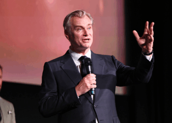 A man in a suit speaks into a microphone on stage, gesturing with one hand. Another man in a suit stands blurred in the background.