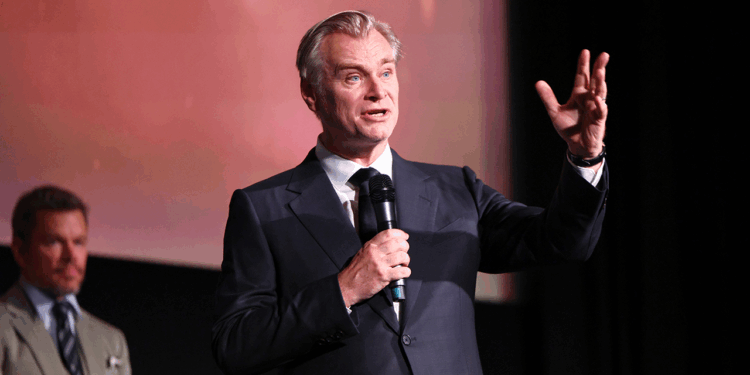 A man in a suit speaks into a microphone on stage, gesturing with one hand. Another man in a suit stands blurred in the background.