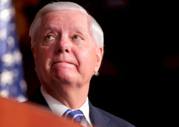 Close-up, low angle shot of Lindsey Graham looking off to his right, with a flag behind him.
