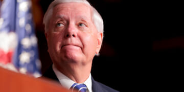 Close-up, low angle shot of Lindsey Graham looking off to his right, with a flag behind him.