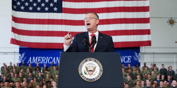 A man in a suit speaks at a podium with the U.S. Vice Presidential seal. An American flag and a group of uniformed military personnel are visible in the background.