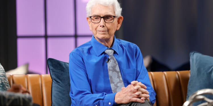 Walt Heyer in a blue shirt and light blue tie sits with his hands folded on the set of a TV show.