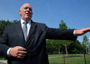 A man in a dark suit stands outdoors, gesturing with his right arm extended to the side, with green trees and a blue sky in the background.