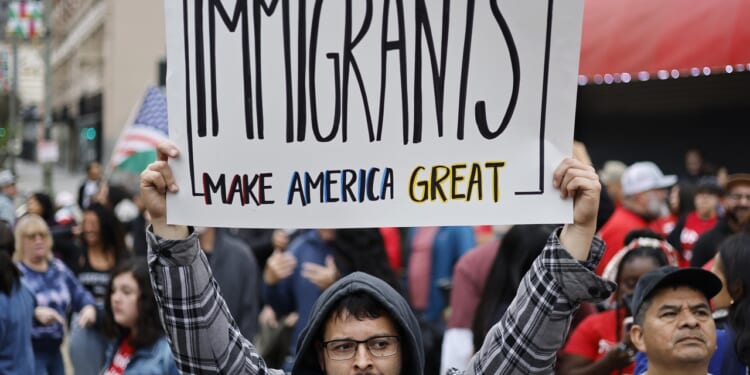 A person in a crowd holds a sign reading Immigrants make America great. People are gathered in the background, some holding flags, at an outdoor event or protest.
