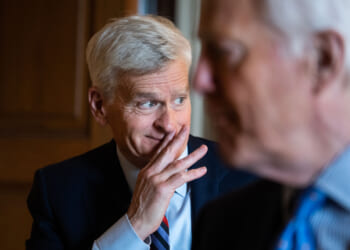 A man with short gray hair in a suit smiles with his hand near his mouth, while another man in a suit is blurred in the foreground. They appear to be having a conversation indoors.