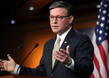 A man in a suit and tie speaks at a podium with microphones. An American flag is in the background. He gestures with his hands, appearing to address an audience or press conference.