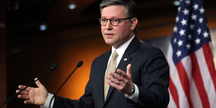A man in a suit and tie speaks at a podium with microphones. An American flag is in the background. He gestures with his hands, appearing to address an audience or press conference.