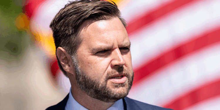 JD Vance, a man with short brown hair and a beard, wearing a suit and tie, stands outdoors in front of a blurred American flag.