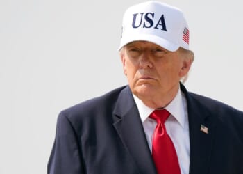 President Donald Trump wears a dark suit, red tie, and a white USA hat standing outside against a plain, light background.
