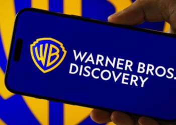 A phone with the Warner Brothers Discovery logo is placed above a blurred screen of the same logo.