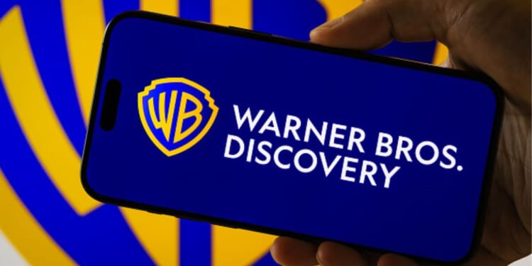 A phone with the Warner Brothers Discovery logo is placed above a blurred screen of the same logo.