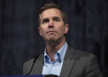 A man in a gray suit and blue shirt stands at a microphone, looking slightly upward. The background is dark and out of focus.