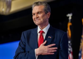Rick Jackson, in a navy suit with a red tie, stands with his hand over his heart, smiling. Two U.S. flags are visible in the background.