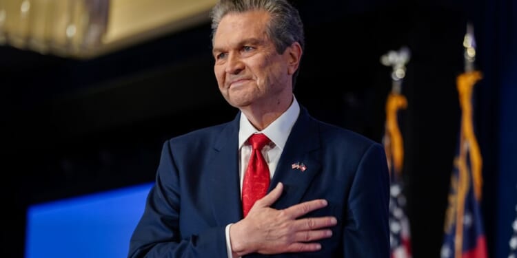 Rick Jackson, in a navy suit with a red tie, stands with his hand over his heart, smiling. Two U.S. flags are visible in the background.