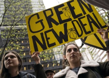 Two people stand outdoors holding a large yellow sign reading “Green New Deal” at a protest, with tall buildings and leafless trees visible in the background.