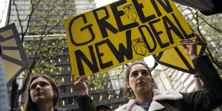 Two people stand outdoors holding a large yellow sign reading “Green New Deal” at a protest, with tall buildings and leafless trees visible in the background.