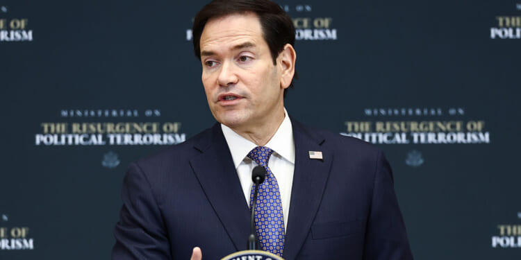 Marco Rubio, wearing a suit and patterned blue tie, speaks at a podium with a sign reading Ministerial on the Resurgence of Political Terrorism and a Department of State seal in front of a dark backdrop. He spoke about leftist terrorism.