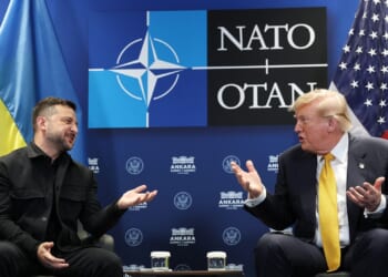 Two men in suits sit facing each other, gesturing while talking. Behind them are Ukrainian, NATO, and U.S. flags, along with a NATO OTAN logo.