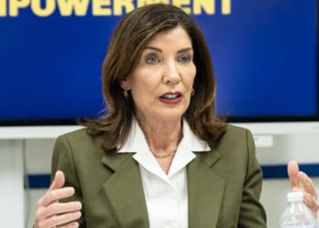 A woman with shoulder-length brown hair wearing an olive blazer and white blouse gestures while speaking, seated at a table with a water bottle. A blurred blue and yellow sign is behind her.