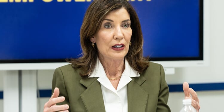 A woman with shoulder-length brown hair wearing an olive blazer and white blouse gestures while speaking, seated at a table with a water bottle. A blurred blue and yellow sign is behind her.