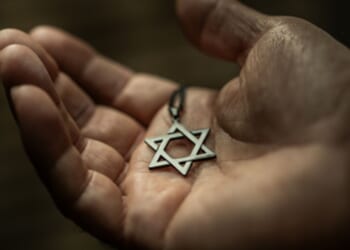 A Star of David necklace sits in a man’s palm in a dimly lit room above a wooden table.