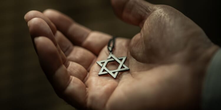 A Star of David necklace sits in a man’s palm in a dimly lit room above a wooden table.