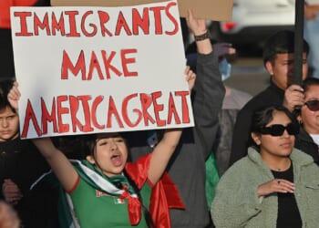 A woman in a crowd holds a sign that reads “Immigrants Make America Great.” Other people, including children and adults, stand around her, some clapping and wearing sunglasses or jackets.