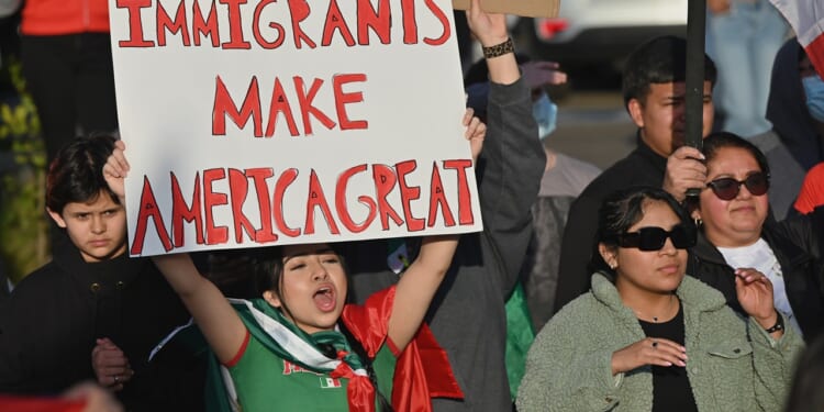 A woman in a crowd holds a sign that reads “Immigrants Make America Great.” Other people, including children and adults, stand around her, some clapping and wearing sunglasses or jackets.