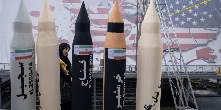 An Iranian woman walks behind mock-ups of four Iranian missiles against a backdrop of an drawing of a scowling Donald Trump.