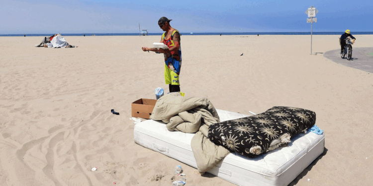 A person stands on a beach next to a pile of bedding and a box. There is a mattress, pillow, and blanket on the sand. Another person rides a bike in the background. The sky is clear and blue.