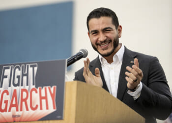 Abdul El-Sayed in a suit smiles and claps at a podium with a sign that reads FIGHT OLIGARCHY.