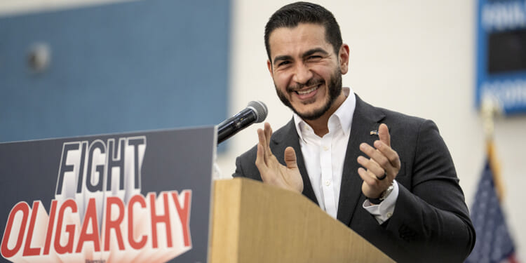 Abdul El-Sayed in a suit smiles and claps at a podium with a sign that reads FIGHT OLIGARCHY.