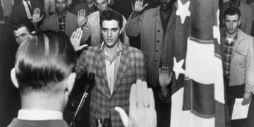 Elvis Presley stands with a group of young men at an induction center, raising their right hands as they are sworn into the United States Army by an officer standing next to an American flag.