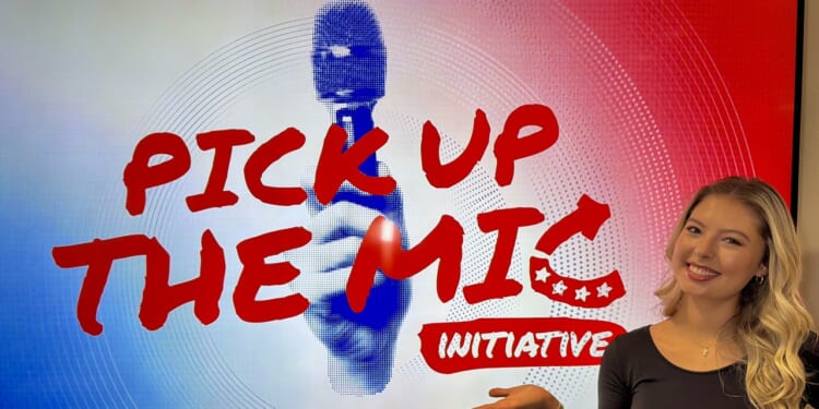 A woman smiles and gestures toward a screen displaying “PICK UP THE MIC INITIATIVE” in bold red letters with a graphic of a hand holding a microphone in the background.
