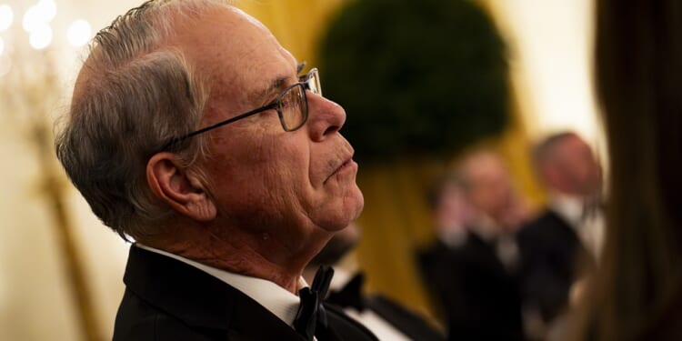 An older man wearing glasses and a tuxedo listens attentively at a formal event, with other people in tuxedos blurred in the background.