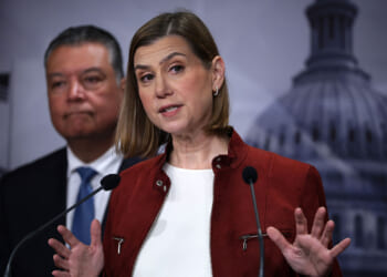 Sen. Elissa Slotkin says the SAVE Act would make it harder for "any Democrat in any state to win any election." A woman in a red jacket speaks at a podium with a man in a suit standing behind her. The blurred background includes a large government building dome and a U.S. flag.