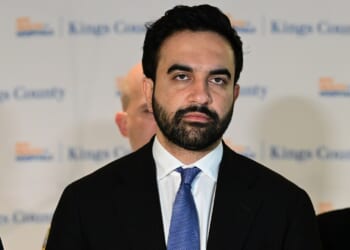 Zohran Mamdani in a suit and blue tie stands in front of a backdrop with blurred text.