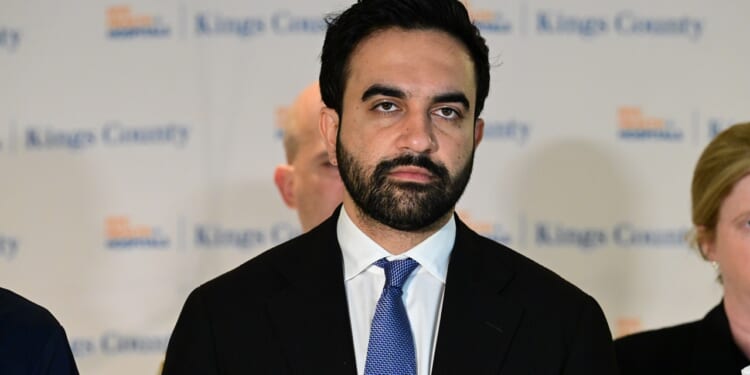 Zohran Mamdani in a suit and blue tie stands in front of a backdrop with blurred text.