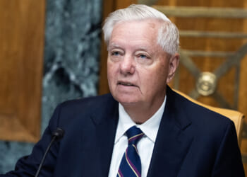 An older man in a suit and striped tie sits at a desk with a microphone, appearing to speak or listen during a formal meeting in a wood-paneled room.