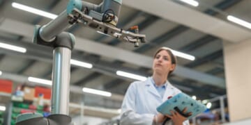 A woman stares at an engineering arm of a machine in a brightly facility.