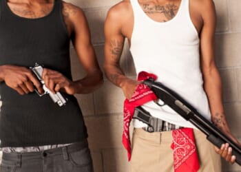 Two gang members stand next to each other with guns just near a brick wall.