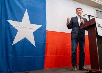 A man stands on stage speaking into a microphone in front of a large Texas flag. A sign reading Talarico for Texas is visible beside him.