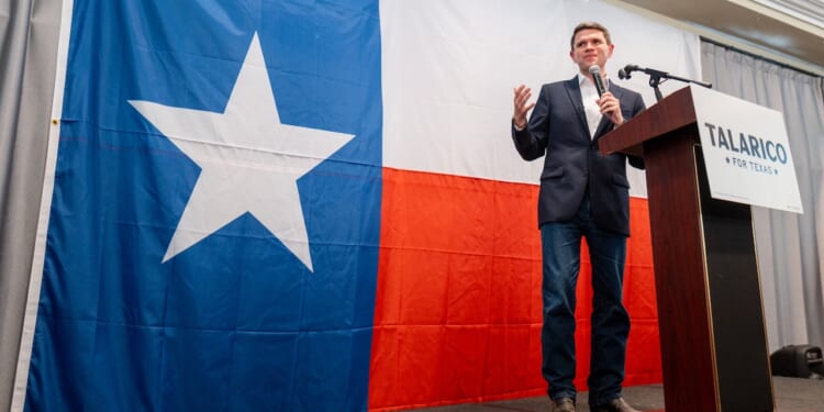 A man stands on stage speaking into a microphone in front of a large Texas flag. A sign reading Talarico for Texas is visible beside him.