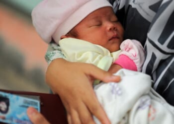 A newborn baby wearing a pink hat and mittens is sleeping in someone's arms. The person is holding a wallet with an ID card visible.