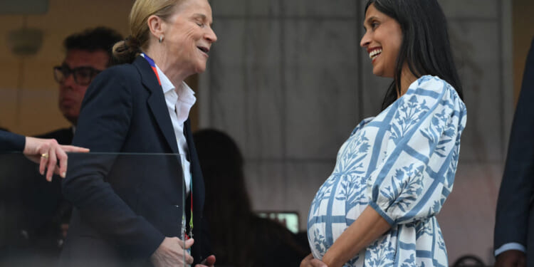 A very pregnant Usha Vance in a white and blue dress speaks to someone at a World Cup match.