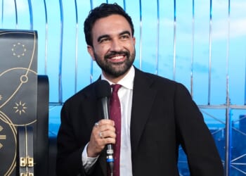 Zohran Mamdani in a suit and tie smiles while holding a microphone. He stands in front of a decorative background with gold designs and metal railings, with a cityscape visible behind him.