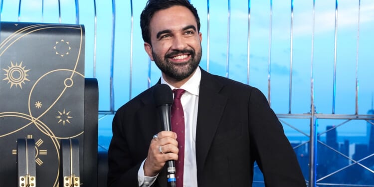 Zohran Mamdani in a suit and tie smiles while holding a microphone. He stands in front of a decorative background with gold designs and metal railings, with a cityscape visible behind him.
